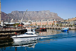 Eine Yacht fährt durch den Hafen von Kapstadt. Im Hintergrund ist der Tafelberg vor blauem Himmel zu sehen